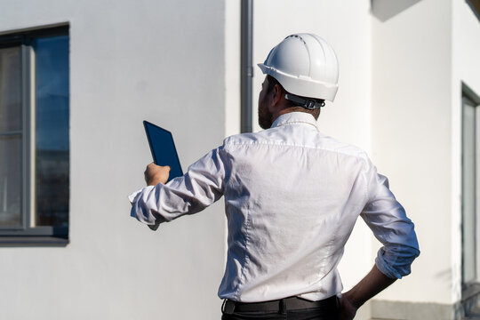 Rear view of foreman in hard hat holding tablet up facing white house wall