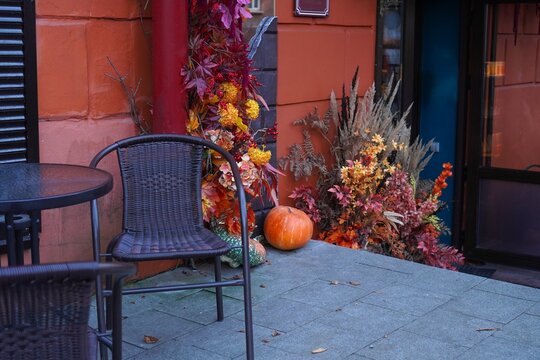 Autumn cafe terrace with wicker chairs, table and pumpkins near decorative red leaves, berries and dried flowers. Seasonal fall decor of outdoor city space for Halloween and Thanksgiving.