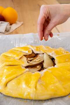 Fruit pastry. Cooking pie. Female baker hand adding pine nuts in pear galette with yellow egg wash on baking sheet at home kitchen.
