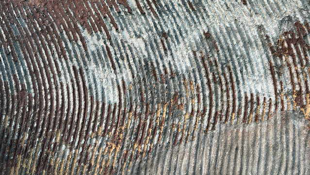 Close-up of a weathered textured surface featuring curved parallel grooves, peeling layers, and earthy color variations, creating a striking abstract pattern and aged background.