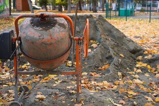 Orange cement mixer with fresh concrete near dug trench in autumn park. Seasonal repair and construction of walkways among fallen yellow leaves, municipal landscaping work in October.
