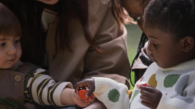 Caucasian child and African American toddler sharing healthy snacks outdoors with mother. Clip suits daycare advertising, parenting content, and child nutrition campaigns