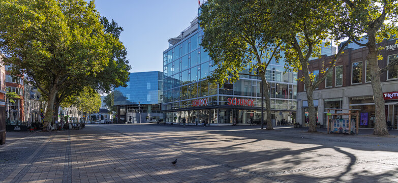 Heerlen, Netherlands - 04 October 2026: Wide perspective of a spacious town square with green trees, modern commercial buildings including SCHUNCK, and shops under a bright blue sky.