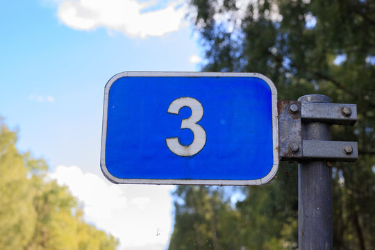 Blue rectangular metal road sign displaying white number three stands mounted on metal pole outdoors. Navigation pointer, route numbering, and traffic direction mark