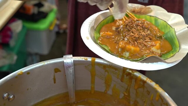 Hand pouring crispy fried shallots onto satay padang lontong with curry sauce