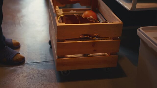 Woman pulling out wooden crate on wheels in kitchen, taking fresh onion and garlic, then rolling the crate back. Smart home storage, cooking preparation, and organization routine.