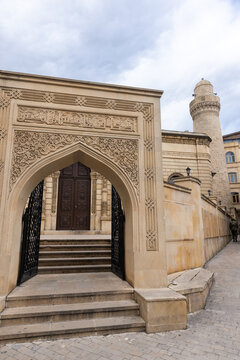 An ornate stone mosque entrance with an arched doorway and intricate carved facade leading up steps to a decorated wooden door, framed by a tall minaret and soft cloudy sky, Baku, Azerbaijan