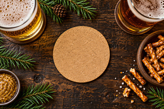 Top view of beer glasses and pretzels on dark wooden table with pine branches and cork coaster, festive snack arrangement. Pub food, hospitality, winter holiday, celebration.