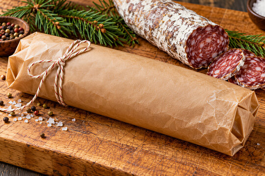 Salami sausage slices and whole cured meat wrapped in brown paper on wooden cutting board, pine branches, spices. Charcuterie, deli, gourmet food, catering.