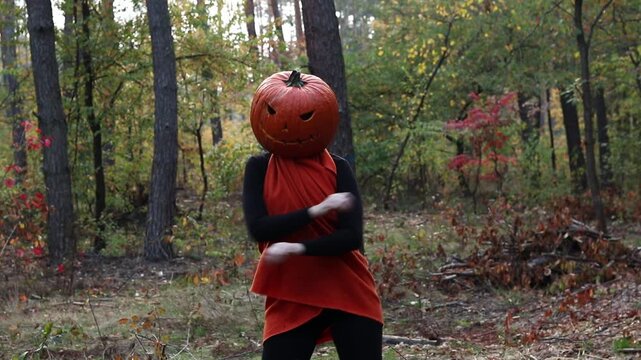 Character with a carved Jack-o-lantern on her head poses and dances in an autumn pine forest. Halloween celebration concept. Girl with a Halloween pumpkin instead of a head. Holiday