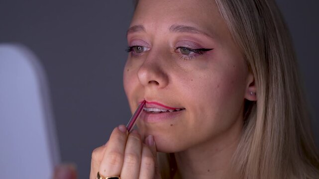 Young blonde woman outlines her lips with a red lip pencil while looking in the mirror, carefully defining the contour in a beauty routine