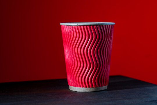 Red corrugated paper coffee cup on dark wooden table. Closeup of red rippled disposable paper cup for hot drinks. Takeaway coffee cup on dark wood desk with solid red background for cafe marketing