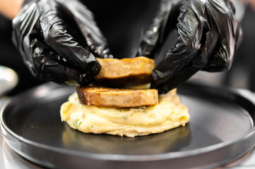 A chef wearing black gloves carefully places a thick, seared slice of foie gras or pork belly onto a smooth bed of mashed potatoes on a sleek black plate, preparing a gourmet culinary dish © pavel siamionov