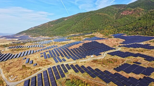 Solar panels cover a large area on a hillside in a rural setting. The rows of panels align with the landscape under a clear sky. Green mountains rise in the background.