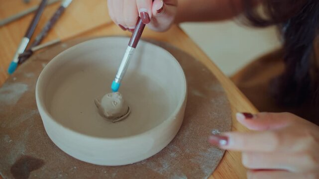 Close up of female artist using paintbrush to prepare clay surface for attaching handmade cat face decoration inside ceramic bowl, creative pottery making process in craft workshop.