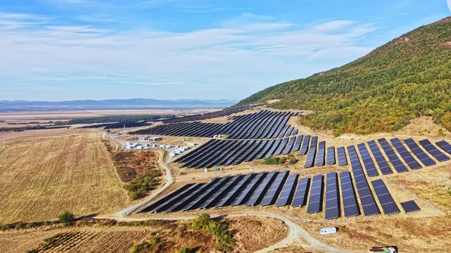 A solar energy farm shows rows of solar panels set on flat land near a hill. The scene captures the arrangement of panels in a rural area under a blue sky.