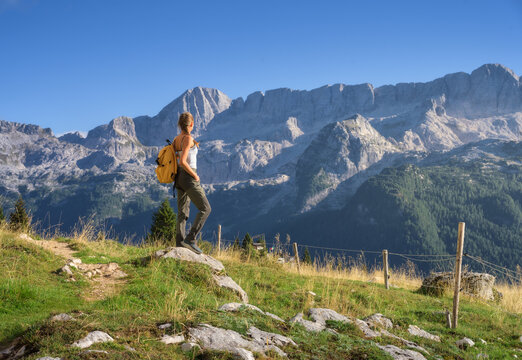 Woman hiker with backpack standing on trail looking at mountains in Slovenia. Alpine autumn landscape with sporty girl, rocky mountain range, blue sky, green grass, pathway and sunny outdoor view