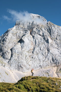 Woman hiker with backpack standing on trail, looking at majestic Mangart mountain in Slovenia. Alpine autumn landscape with girl, huge rocky peak, blue sky, clouds, green grass and sunny day. Hiking