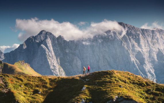 Two women hikers walking along a grassy mountain ridge, rocky peaks in clouds in the Julian Alps, Slovenia, with warm evening sunlight illuminating the alpine landscape at sunset in autumn. Nature