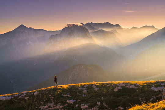 Woman hiker standing on rocky mountain ridge at sunset. Fall in Slovenia. Alpine landscape with sunbeams through fog, rocky peaks, clear sky, low clouds, golden autumn grass and dramatic evening light