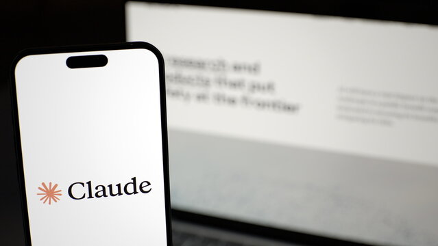 The Claude logo displayed on a smartphone held in front of a laptop. Anthropic commits $100 million to train 10,000 AI engineers by 2027. October 4, 2026, Lisbon, Portugal.