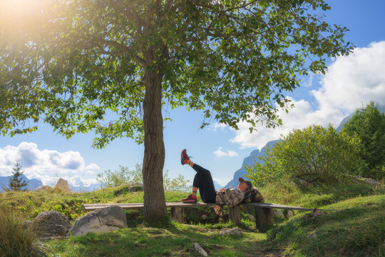 Woman hiker lying down on wooden bench resting under green tree in Slovenia. Alpine landscape with girl, mountains, blue sky, clouds, green grass meadow, backpack and sunny outdoor view in summer