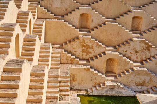 Ancient Indian stepwell featuring symmetrical geometric staircases descending into the earth, constructed from beige sandstone. The architectural marvel displays multiple tiers of zigzagging steps