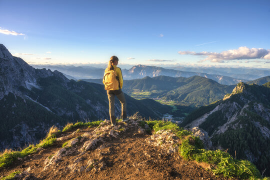 Woman hiker with yellow backpack standing on rocky mountain summit in Slovenia. Alpine autumn landscape with panoramic view of peaks, sky, clouds, green hills, sun flare and evening sunset light