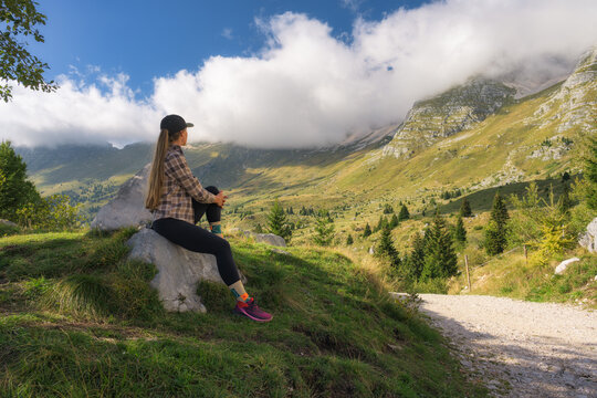 Woman hiker sitting on rock near trail looking at mountains in Slovenia. Alpine autumn landscape with girl, green grass meadow, trees, cloudy sky, dirt road and sunny outdoor scenery in fall
