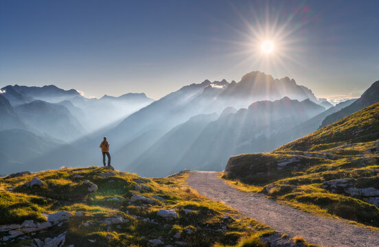 Woman hiker with backpack standing on trail in Slovenia. Alpine mountain landscape with sunstar, sunbeams through fog, sky, path, autumn grass and golden light. Silhouette of woman and mountain peaks