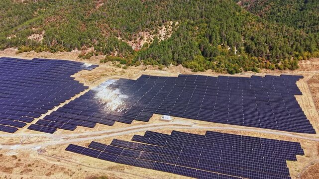 The solar panel farm contains many rows of black solar panels arranged on the ground. The area is surrounded by trees and hills, with no buildings nearby. Sunlight reflects off the panels.