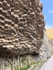 Textured basalt columns form a towering cliff, with moss at its base and a paved walkway bordered by posts beneath a bright blue sky © Anat art