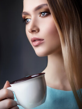 A young woman with blonde hair holds a white mug with a dark liquid in it near her mouth. She is looking away from the camera.