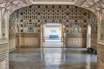 An ornate interior chamber within a historical Indian palace, featuring intricately decorated walls with colorful vase and bottle motifs inlaid with mirrors and glass mosaics. The polished marble © Andrius