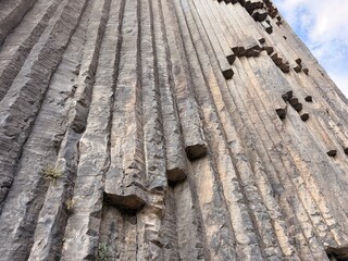 Close view of a tall gray basalt cliff showing vertical columns, rugged weathering, scattered plants, protruding rocks, and sky above © Anat art