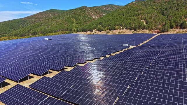 A vast field of solar panels is set in a rural area. The panels are aligned in rows, reflecting sunlight. Mountains and trees provide a natural backdrop.