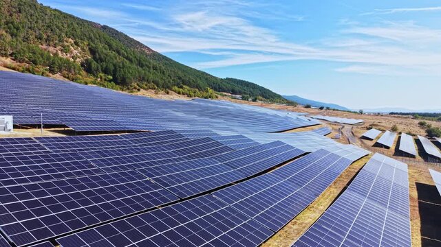 Rows of solar panels are set up in a field with a backdrop of green hills on a sunny day. The area is focused on generating clean energy.