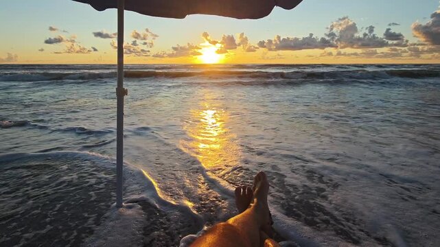 Personal perspective of a man's feet relaxing on the wet sand under an umbrella as gentle ocean waves wash ashore during a beautiful and serene golden sunset, symbolizing tranquility and vacation