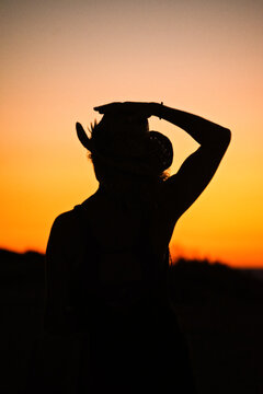 A woman seen from behind in backlit holding a Texas cowboy hat, silhouette in backlit at sunset. Book cover, symbolic, poster, chasing sunset 

