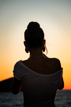 A woman seen from behind in backlit with a messy bun, dangling earrings and a white dress watching sunset. Silhouette in backlit at sunset. Book cover, symbolic, poster, chasing sunset 
