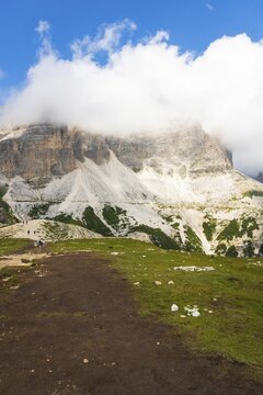 Scenic dirt hiking path through green alpine meadow towering limestone cliffs mountain peak under fluffy white clouds background Sella Pass Dolomites Italy summer landscape