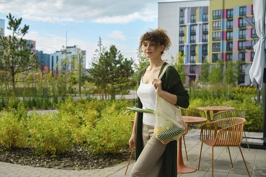 Cute woman returns home from farm store carrying fresh vegetables and fruits in reusable mesh bag. She goes through landscaped green area in residential area with apartment buildings