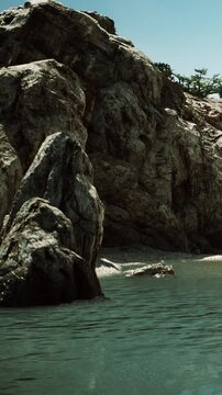 Rocks stand near calm water under a clear blue sky in a coastal area. The scene shows textured stone surfaces and reflections on the water during daytime.