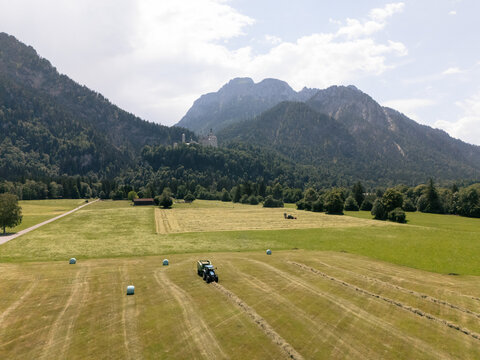 Aerial view of a tractor working in a hay field with hay bales, surrounded by lush green meadows and the dramatic peaks of the Tiroler Zugspitz Arena in Tirol, Austria.