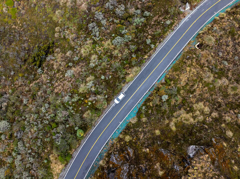 Aerial view of a car driving along a winding road through the rugged, shrub-covered landscape of the Nevado del Ruiz region in Villamaria, Caldas, Colombia.