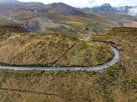 Aerial view of a winding mountain road traversing the rugged paramo landscape near Nevado del Ruiz, with a yellow bus and a car driving along the curve in Villamaria, Caldas, Colombia.