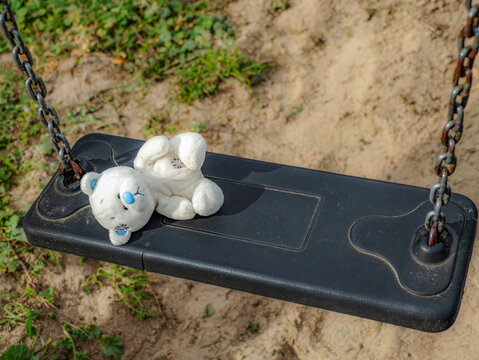 Lonely White Teddy Bear Left On Empty Playground Swing Suggesting Childhood Loss And Nostalgia