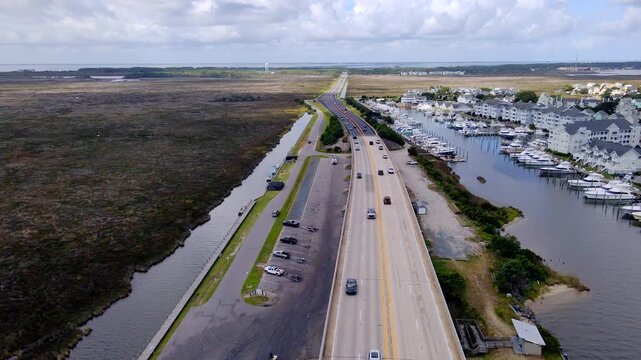 Cars travel along a coastal road between broad marshland and the marina waterfront.