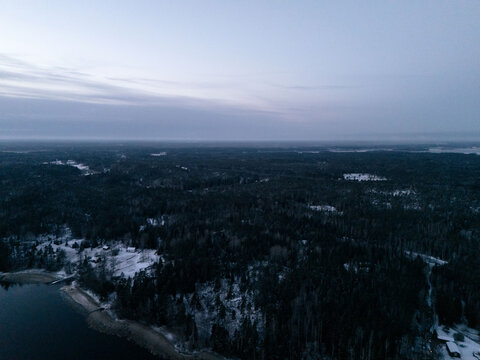 Aerial view of a vast, snow-covered forest landscape bordering a dark lake under a dim twilight sky in Uusimaa, Finland.