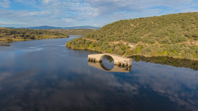 Aerial view of the Roman bridge of Caparra partially submerged in the Gabriel y Galan reservoir with calm water and surrounding hills in Guijo de Granadilla, Extremadura, Spain.